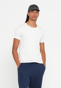 Fiatal férfi hosszú fonatokkal, fekete sapkában, fehér, rövid ujjú pólóban és tengerészkék nadrágban áll, kezét a zsebében tartva.