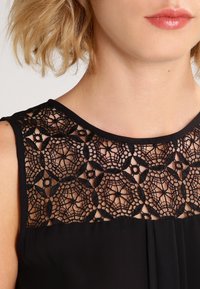 Robe noire avec un empiècement en dentelle texturée au niveau de l'encolure, présentant des motifs circulaires, un tissu contrastant en dessous, et une coupe épurée et ajustée.