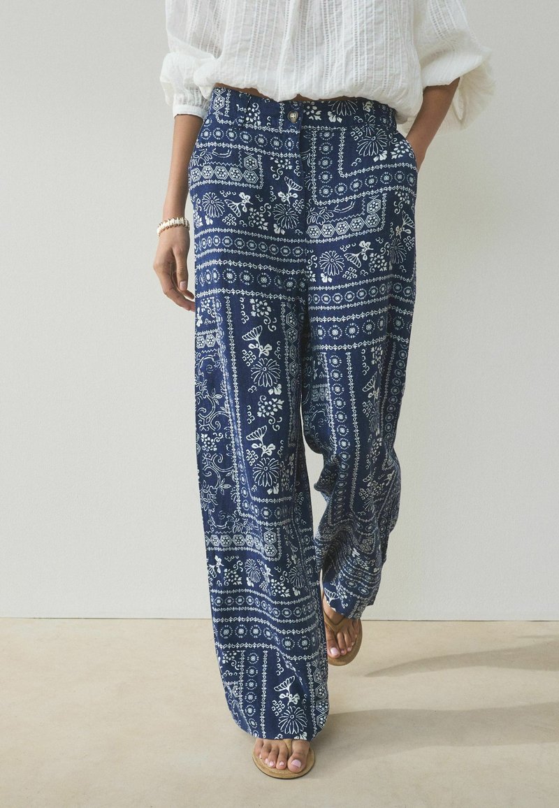 Femme portant un pantalon bleu marine ample avec des motifs floraux et géométriques blancs, associé à un chemisier blanc et des sandales.