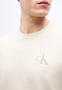 Εγγύς πλάνο ενός ατόμου που φοράει μια μπεζ μπλούζα με ένα μικρό ανοιχτό γκρι λογότυπο "CK Calvin Klein" στην αριστερή πλευρά του στήθους.