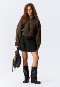 Ylivoimainen ruskea huppari vetoketjulla, musta laskostettu mini-hame, mustat polvipituiset saappaat ja pehmeä, kerätty musta laukku, jossa on turkisakseli.