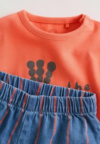 Narančasta pamučna majica s grafičkim dizajnom crnih oblika, u kombinaciji s plavim traper hlačicama s crvenim okomitim prugama i elastičnim pojasom.