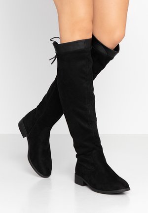 Bottes en daim noir montant jusqu'au genou avec des talons blocs bas, portées sur des jambes croisées sur un fond clair uni.
