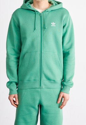 Mężczyzna ubrany w turkusową kurtkę na zamek marki Adidas z kieszeniami kangurzymi oraz dopasowane spodnie dresowe na białym tle.