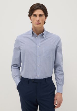 Uomo con camicia a quadri azzurra chiaro abbottonata e infilata in pantaloni blu scuro, in piedi con una mano in tasca davanti a uno sfondo semplice.