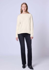 Cremfarbener Strickpullover mit langen Ärmeln, rundem Ausschnitt und lockerer Passform, kombiniert mit dunklen Jeans und Ankle Boots. Glatte Textur, minimales Design.