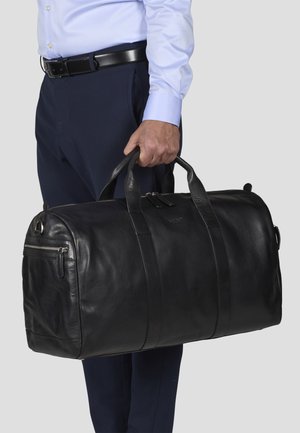 Bolsa de viaje de cuero negro con asas enrolladas, que cuenta con una parte superior con cremallera y un bolsillo frontal. Textura suave y detalle de costura visible.