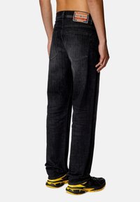 Jeans de mezclilla negros con corte recto, que cuentan con bolsillos traseros y una etiqueta de cuero con marca. Usados con zapatillas deportivas negras y amarillas.