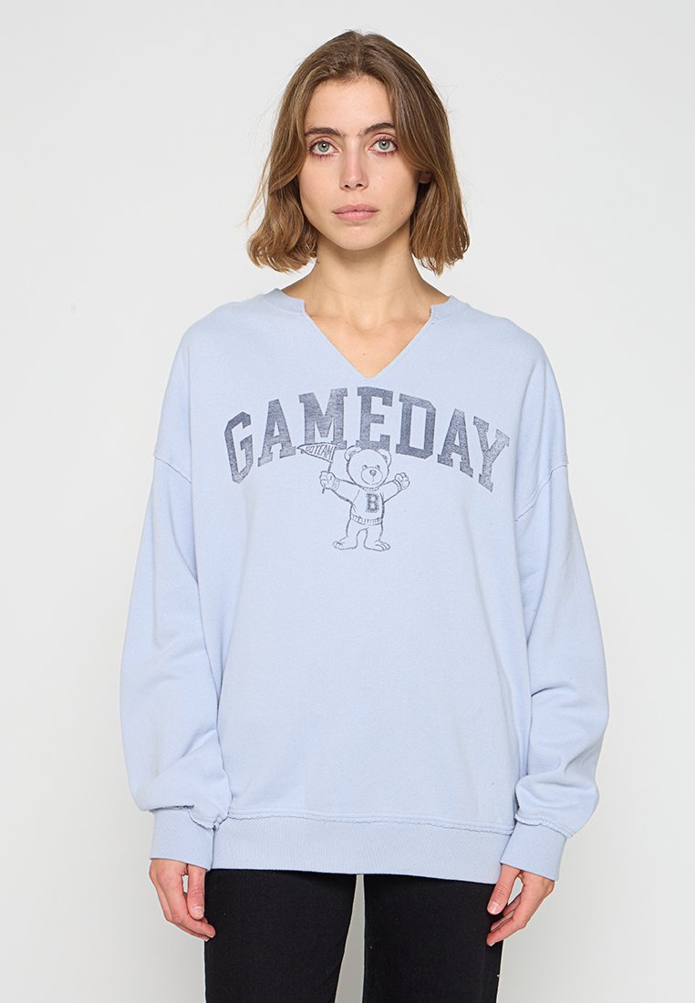 Hellblauer, übergroßer Hoodie mit V-Ausschnitt, auf dem der Text "GAMEDAY" und eine graue Cartoonbär-Grafik zu sehen sind. Aus weichcotton Material.