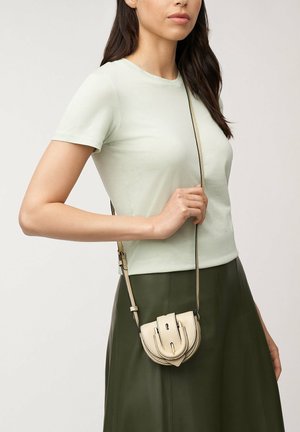 Vrouw draagt een lichtgroen shirt met korte mouwen en een donkergroene rok, en houdt een kleine beige schoudertas met gespdetail vast.