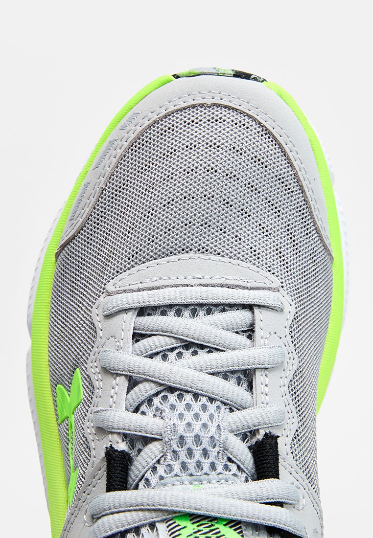 Primer plano del zapato deportivo de malla gris con acentos en verde neón y cordones blancos atados en un patrón de cruz.