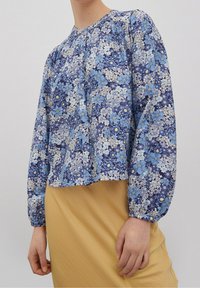 Blusa a maniche lunghe con motivo floreale blu e collo arricciato, abbinata a una gonna gialla realizzata in un tessuto morbido e testurizzato.