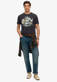 Marine T-shirt med grafisk design, blå jeans, brun bælte og en ternet skjorte bundet om taljen. Beige sneakers fuldender outfittet.