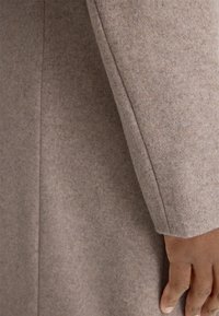 Cappotto in lana marrone, tessuto texture, maniche aderenti con cuciture pulite e finitura liscia. Calore notevole e design strutturato.