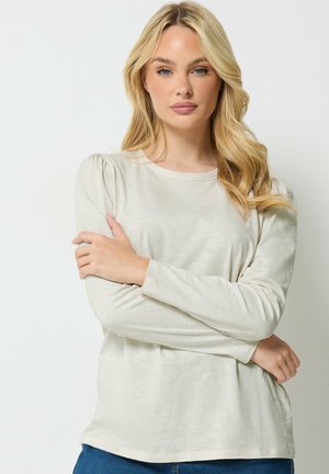 Mujer rubia con rizos sueltos que lleva una camiseta de manga larga beige claro y vaqueros azules, de pie con los brazos cruzados sobre un fondo blanco.