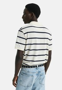 Korte mouw polo in lichtbeige met marineblauwe strepen, met een geribbelde kraag en een ontspannen pasvorm. Draag met lichtblauwe spijkerbroek.
