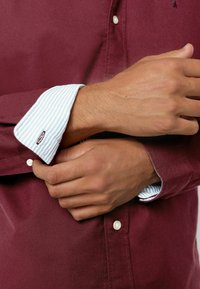 Camisa burdeos con botones, con puños a rayas azul claro en contraste. Presenta botones blancos y una textura suave. Sin patrones visibles.