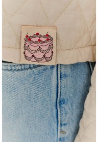 Beżowa tkanina z pikowaniem ma ilustrację różowego tortu warstwowego z czerwonymi wiśniami na wierzchu, przymocowaną do jasnoniebieskiego denimu.