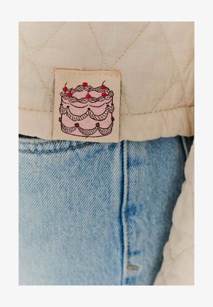 Kvadrovaná béžová látková značka zobrazuje ilustraci vrstveného růžového dortu s červenými třešněmi na vrcholu, připevněná k světle modrému denimu.