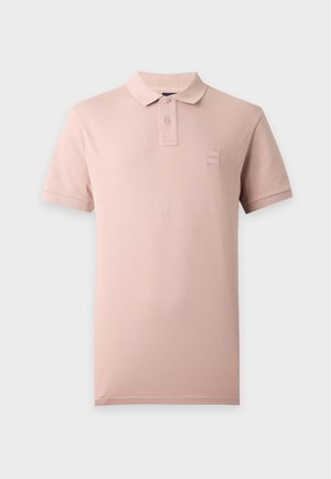 Un mannequin masculin se tient debout, les mains dans les poches, vêtu d’un polo rose pâle, d’un pantalon noir ample et de chaussures foncées sur un fond uni.