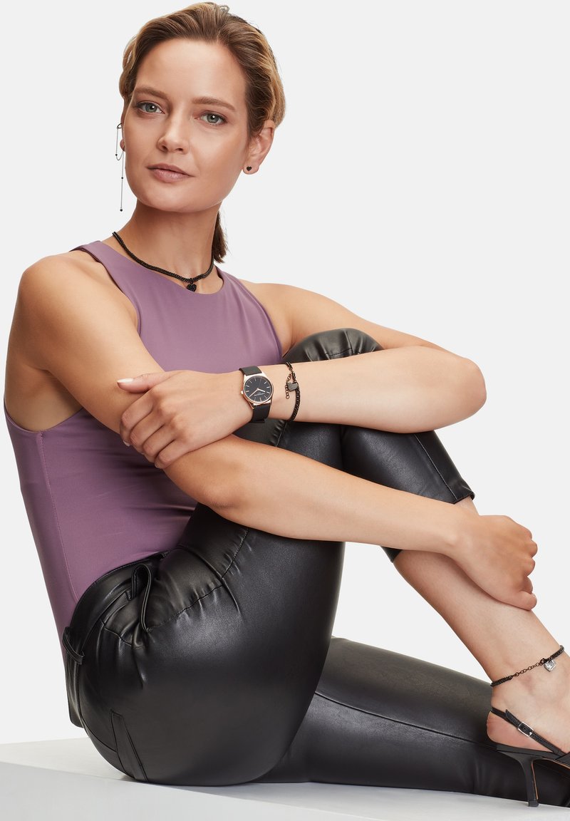 Anliegendes lavendelfarbenes Tanktop, schwarze Leggings aus Faux-Leder und Accessoires, darunter eine Uhr und mehrlagige Halsketten. Sanfte Textur und elegantes Design.