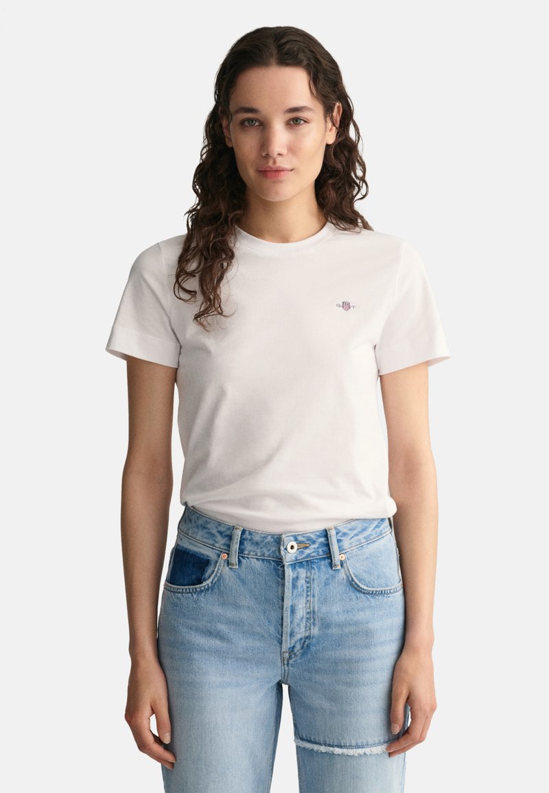 Witte katoenen t-shirt met korte mouwen, ronde halslijn en een klein geborduurd logo op de borst, gecombineerd met lichtblauwe, versleten jeans.