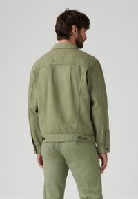 Jachetă de culoare verde măsliniu cu un design structurat, având manșete cu butoane și o margine dreaptă. Jacheta are o textură netedă, mată și cusături discrete.