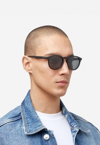 Occhiali da sole rotondi neri con lenti scure, finitura opaca e aste piatte, indossati da una persona con una giacca di jeans blu su uno sfondo semplice.