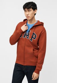 Νέος άνδρας που φοράει φούτερ GAP χρώματος σκουριάς πάνω από μπλε πουκάμισο, ρυθμίζει το φερμουάρ, στέκεται μπροστά σε απλό φωτεινό φόντο.