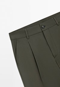 Close-up of olive green tailored trousers showing waistband, belt loops, front pleats, side pocket, and single button closure.