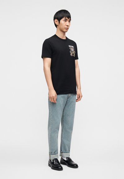 Μαύρο βαμβακερό μπλουζάκι με γραφικό αρκούδας στο αριστερό στήθος, συνδυασμένο με ανοιχτόχρωμο τζιν παντελόνι και μαύρα μοκασίνια.