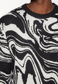 Gros plan sur un motif abstrait tourbillonnant noir et blanc sur un pull en tricot porté par une personne à la peau foncée.