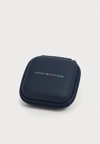 Jūras zilas krāsas, ar rāvējslēdzēju, ar gludu tekstūru, priekšpusē ar modernā fontā rakstītu baltu logo "TOMMY HILFIGER". Kvadrāta forma.