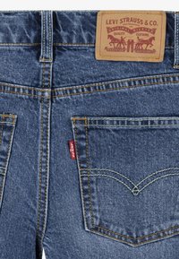 Jeans de mezclilla azul oscuro con costuras amarillas, que presentan un parche de cuero etiquetado "Levi Strauss & Co." y una pequeña etiqueta roja en el bolsillo trasero.