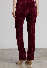Mujer vestida con pantalones rectos de terciopelo rojo oscuro y zapatos de tacón beige, de pie sobre un suelo de concreto.