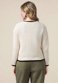 Sweter o kremowej fakturze z czarnym wykończeniem przy kołnierzu i mankietach, zestawiony z oliwkowymi spodniami. Skoncentruj się na wzorze dzianiny i kontrastujących kolorach.