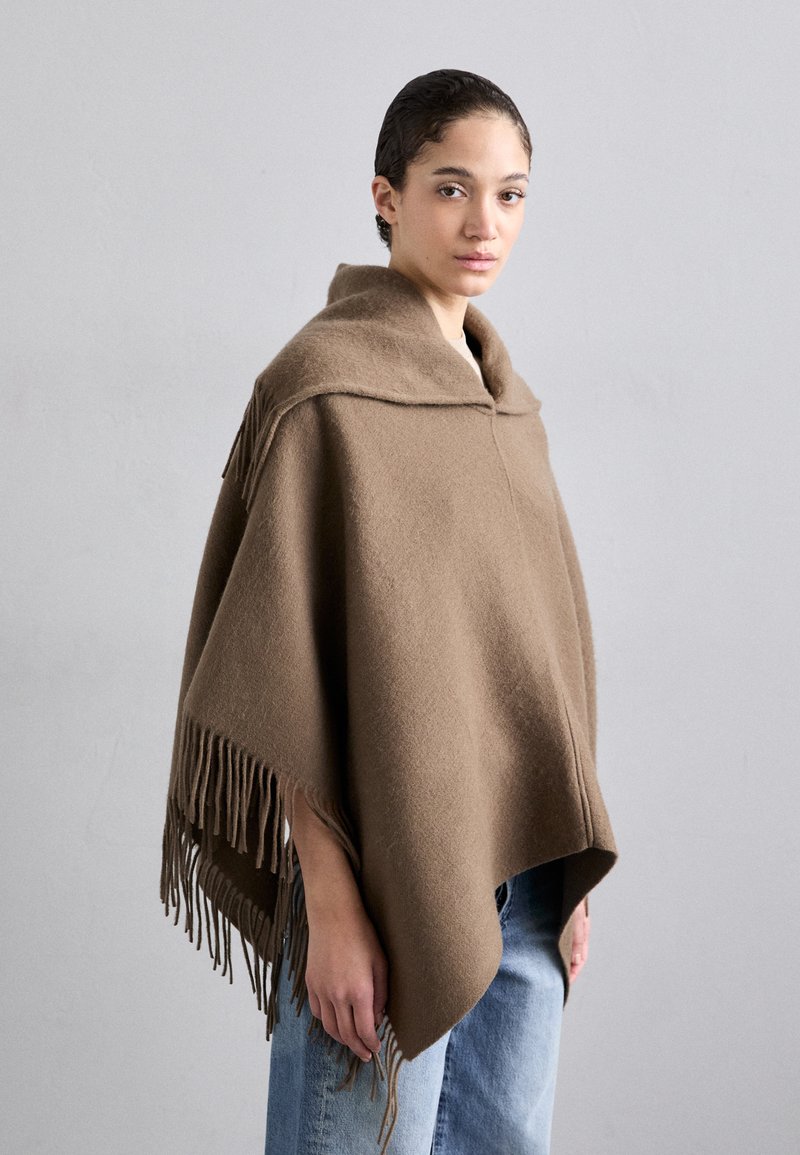 Brūns vilnas pončo ar salocītu apkakli, ar frizētiem detaļām ap hemu un brīvu piegriezumu, uzvilkts virs gaiši zilām džinsām.