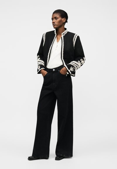 Γυναίκα που φοράει ένα μαύρο σακάκι με λευκές κεντητές λεπτομέρειες πάνω από ένα λευκό πουκάμισο και μαύρο παντελόνι ψηλόμεσο με φαρδιά παντελόνια, όρθια με τα χέρια στις τσέπες.