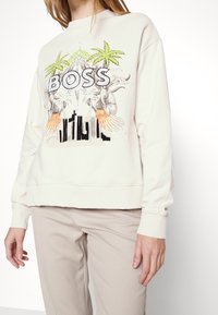 Krémová mikina s potiskem zahrnujícím palmy, slovo "BOSS" a abstraktní vzory. Měkká tkanina s žebrovanými manžetami a lemováním.