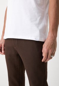 Camiseta blanca de algodón con un dobladillo recto, combinada con pantalones marrones texturizados. Primer plano de la zona de la cadera, que muestra un ajuste casual y detalles de las manos.