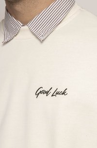 Crewneck sweatshirt i off-white stoff med en kursiv "Good Luck" trykk. Har en brun og hvit stripete krage. Glatt tekstur.