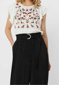 Blusa bianca con ricami floreali in rosso e rosa, abbinata a pantaloni neri a gamba larga con vita belted e un accentuazione in fibbia argentata.