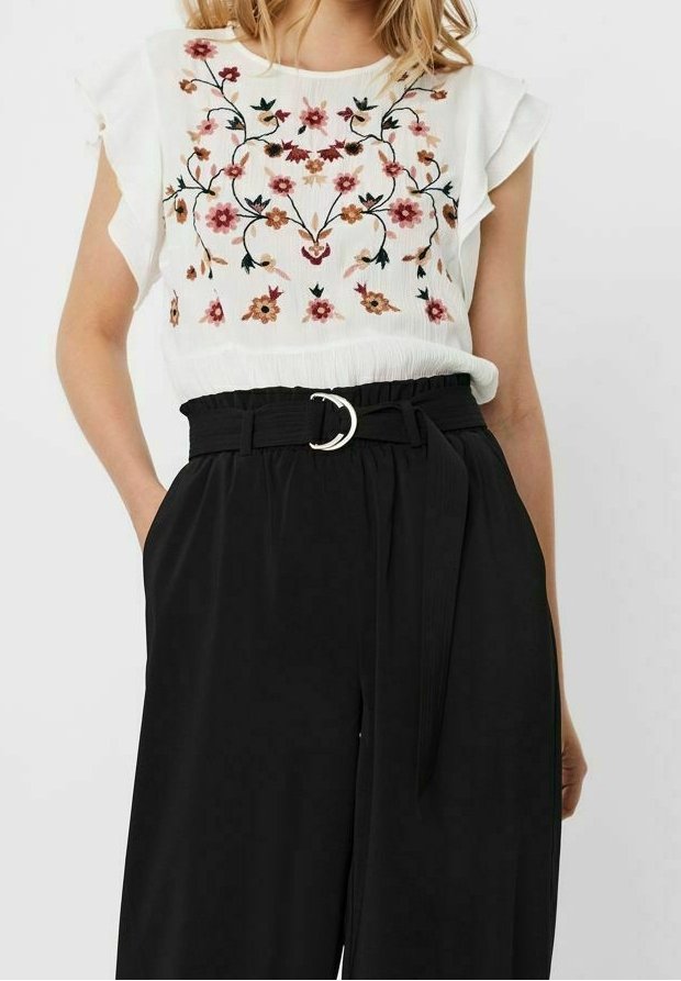 Blusa bianca con ricami floreali in rosso e rosa, abbinata a pantaloni neri a gamba larga con vita belted e un accentuazione in fibbia argentata.