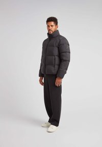 Giacca a vento nera con colletto alto, design trapuntato e chiusura a zip. Presenta un logo sulla manica sinistra. Indossata con pantaloni scuri e scarpe bianche.