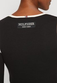 Schwarzes langärmliges Oberteil mit Rippstruktur und einem kontrastierenden cremefarbenen Halsausschnitt. Enthält ein rechteckiges "HILFIGER NEW YORK"-Label auf der Rückseite.