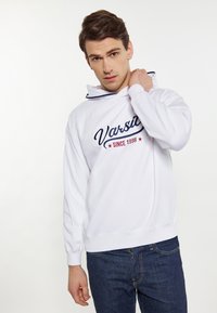 Witte hoodie met marineblauwe borduursels "Varsity" en "SINCE 1998", voorzien van een trekkoord en geribde boorden. Gedirigeerd met blauwe spijkerbroeken.