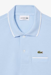 Ljusblå polo med vit kant, treknappsknäppning och en grön broderad krokodil-logga. Tillverkad av textil med struktur.