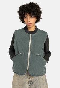 Groen, dik katoenen vest met een afgeronde zoom, zilveren ritssluiting, twee zijzakken en een geribde halslijn. Draagt over een zwart shirt met lange mouwen.