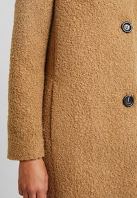 Primo piano di una manica di un cappotto in lana textured beige con bottoni neri e una mano visibile nell'angolo in basso a sinistra.