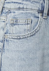 Ljust blå denimjeans har en bakficka med synlig sömnad och en metallnitar, vilket visar en blekt, texturerad yta.